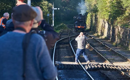 Steam train photographers The photograph, taken in the afternoon during the autumn of 2018, captures a group of steam train photographers at a station on the North Yorkshire Moors in the United Kingdom. The main subject of the image is the enthusiastic observers, some of whom are standing on or near the railway tracks with their cameras aimed at a moving steam train, which is approaching and producing visible smoke. The scene conveys the transport setting of a rural station, with the classic train being a focal point amidst the greenery and stone walls characteristic of the North Yorkshire Moors. This image is best described as a landscape photograph, highlighting the historic appeal of steam trains and train travel in a countryside station environment.